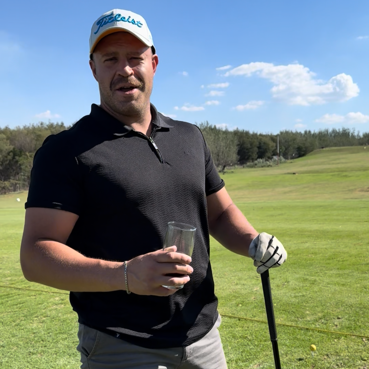 Man on a golf course holding a glass and a golf club with a clear blue sky in the background